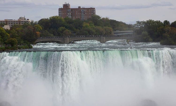 Wide view of Niagara Falls dropping into mist, a bridge above the rushing river, with buildings & trees in the distance.