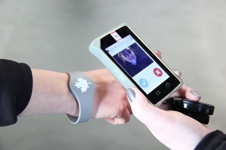 Hand wearing a gray maple leaf wristband near a handheld device displaying a smiling woman's photo and verification icons.