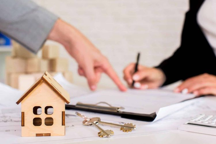 Hands signing a contract with a model house and keys on the table.