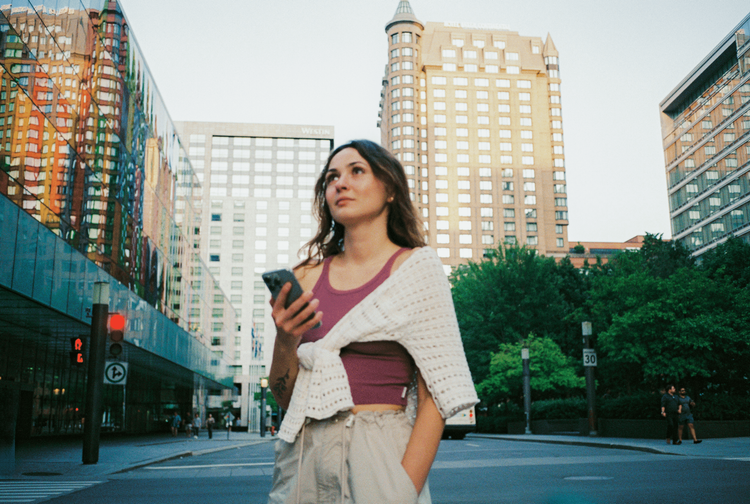 Young woman holding a phone on a city street with tall, reflective buildings and a hotel tower under a light sky.