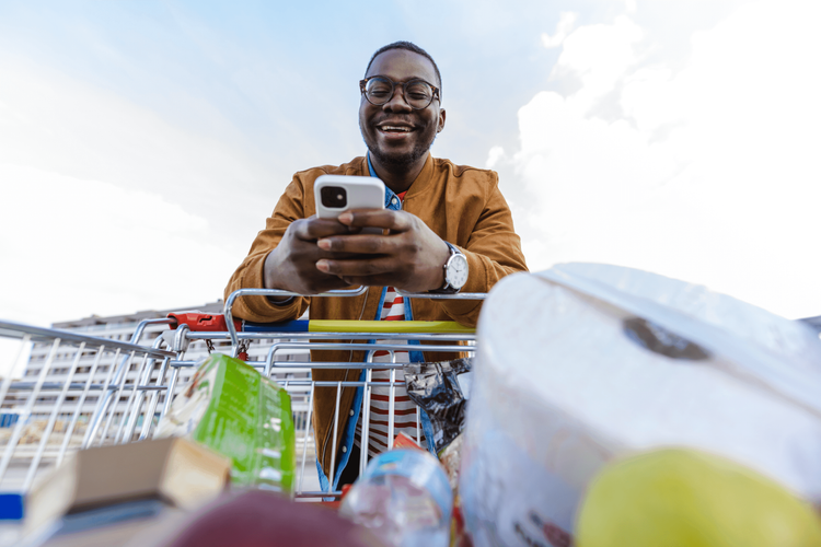 Smiling Black man in glasses checks his phone while pushing a grocery cart filled with items.