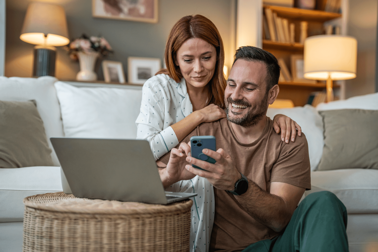 Smiling couple looking at a smartphone together in a cozy living room with a laptop nearby.