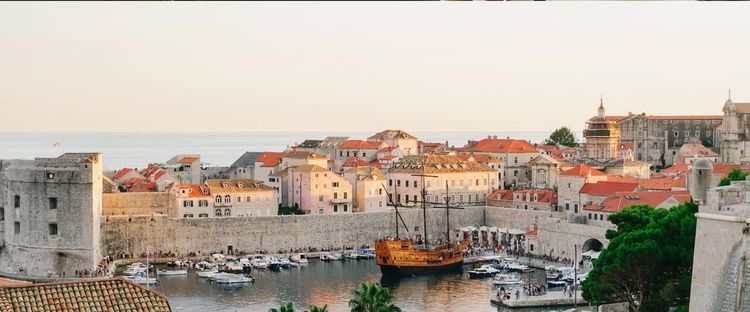 Dubrovnik's walled city panoramic view: harbor with boats, large wooden pirate ship, red-roofed buildings, sea.