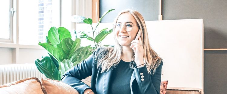 Blonde woman with glasses smiles while talking on a phone in a bright room with a green plant.