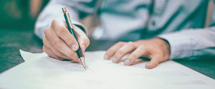 A person's hand holds a pen, writing on a document. Another hand rests on the page.