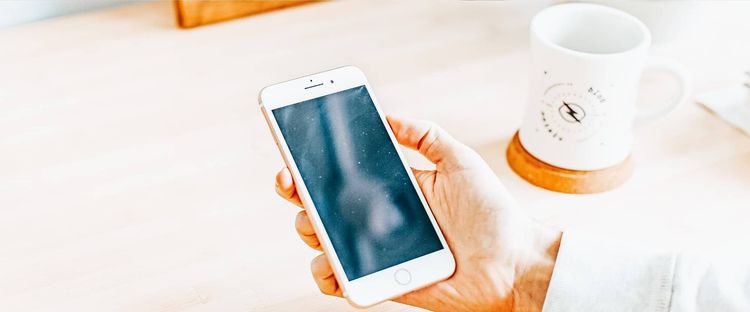 Hand holding white smartphone with dark screen. White mug on wooden coaster in background.