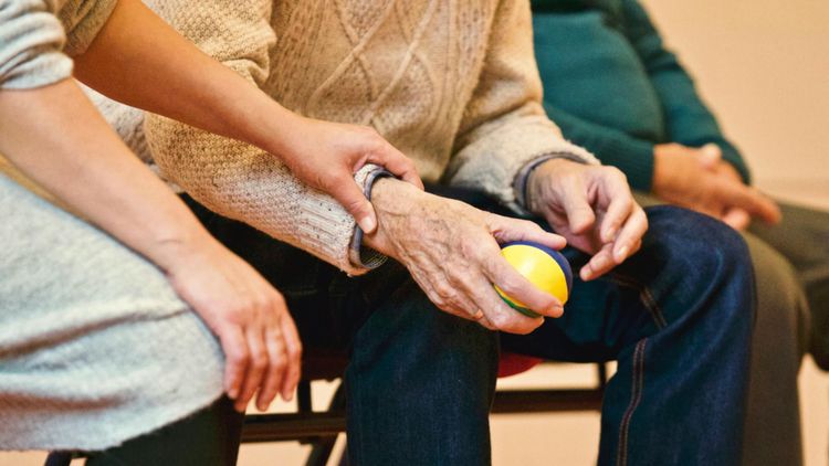 A young person's hand rests on an older adult's arm, who holds a yellow, blue, and green therapy ball.