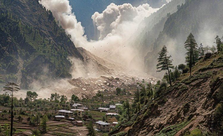 Massive landslide cascades down a mountain, filling a valley with dust clouds above a village with terraced fields.
