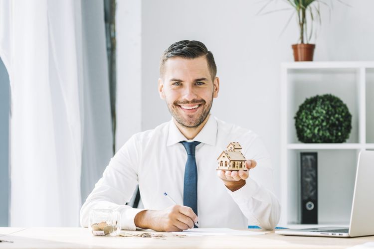 Smiling man in a white shirt and blue tie holding a wooden house model, signing documents at an office desk.