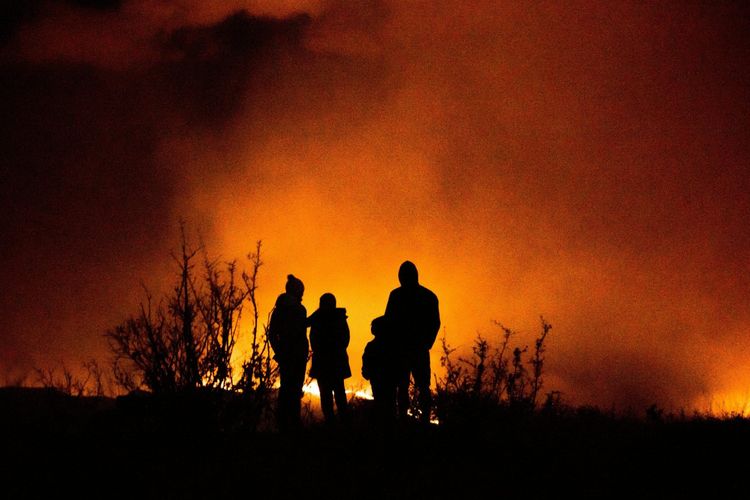 Three people and trees in silhouette against a fiery orange, smoky sky, with flames visible below.