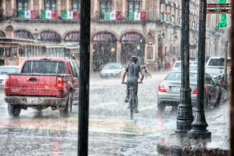 A person bikes through heavy rain on a city street with cars and a building draped in Mexican flags.