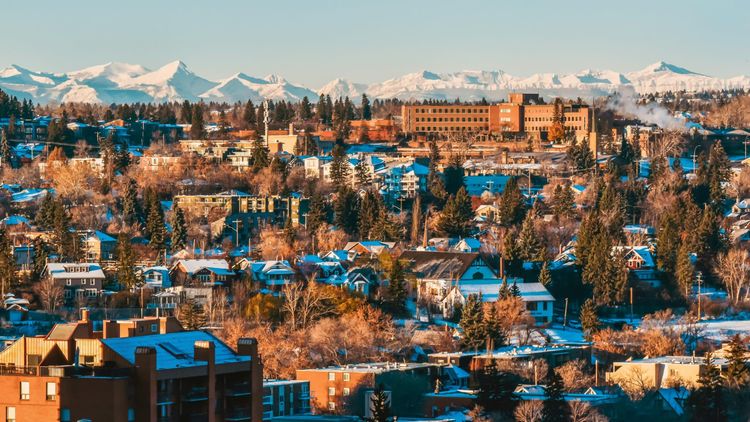 Golden hour illuminates a snowy city nestled among trees, with a prominent brick building and majestic snow-capped mountains.